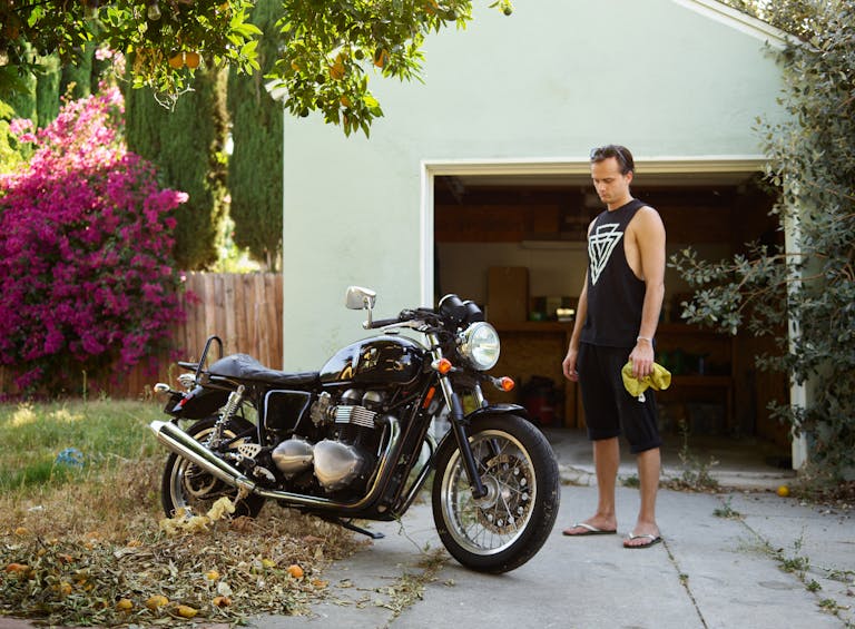 A man with a towel stands beside a motorbike in a suburban driveway.