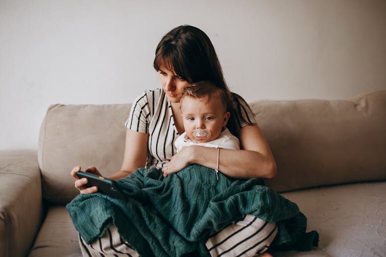 A mother and her baby relax on a cozy sofa, wrapped in a blanket, indoors.