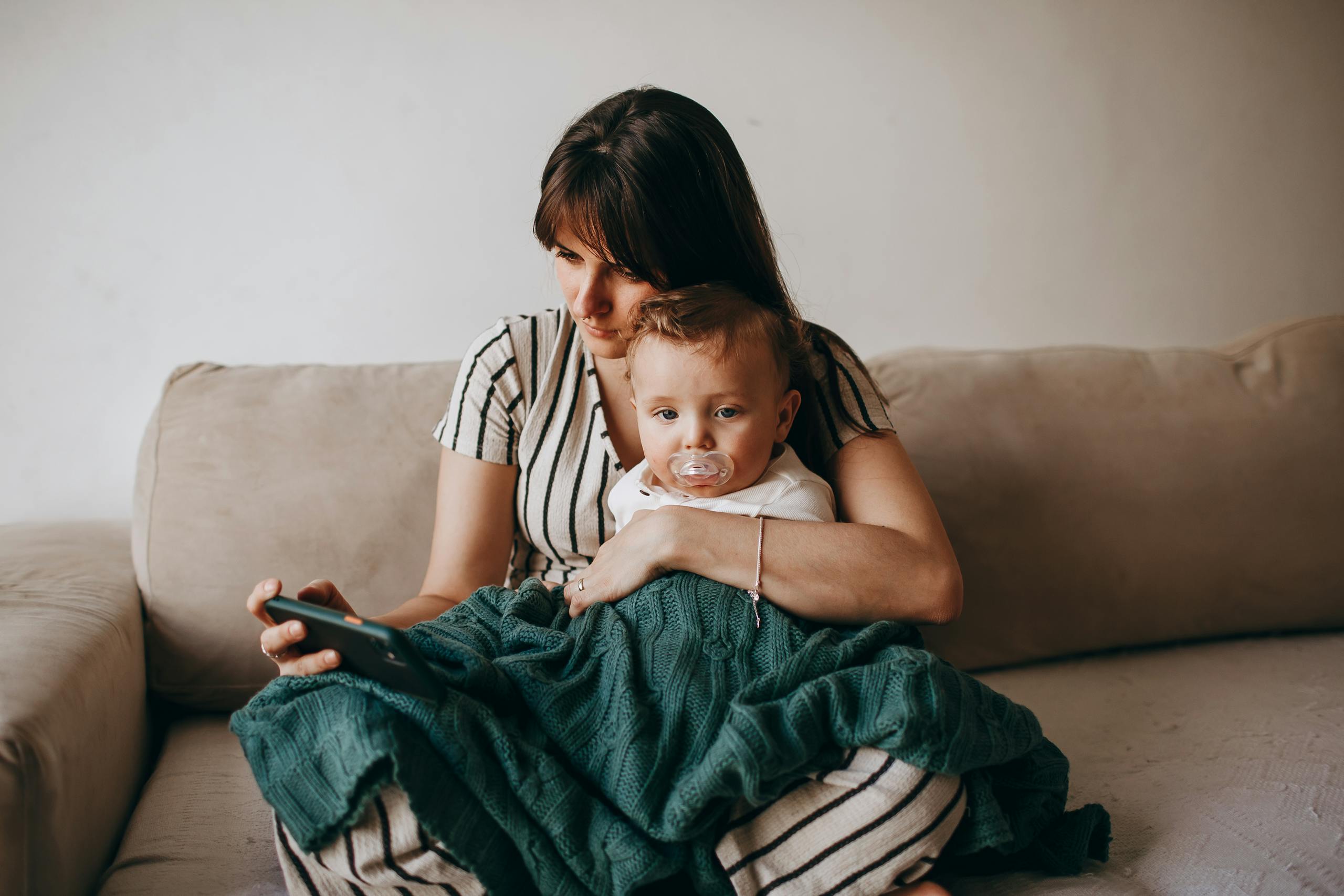 A mother and her baby relax on a cozy sofa, wrapped in a blanket, indoors.