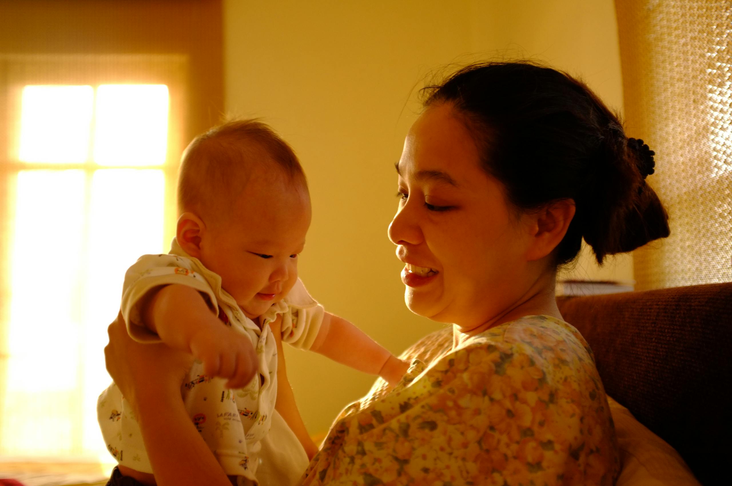 A mother lovingly holds her baby near a sunny window, creating a warm, affectionate moment.