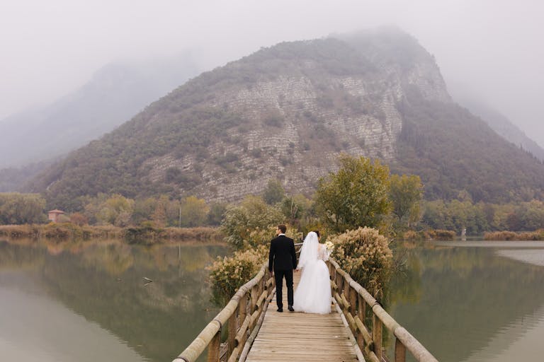 A newlywed couple walks hand in hand on a scenic bridge in Lombardy, Italy, surrounded by serene mountain views.
