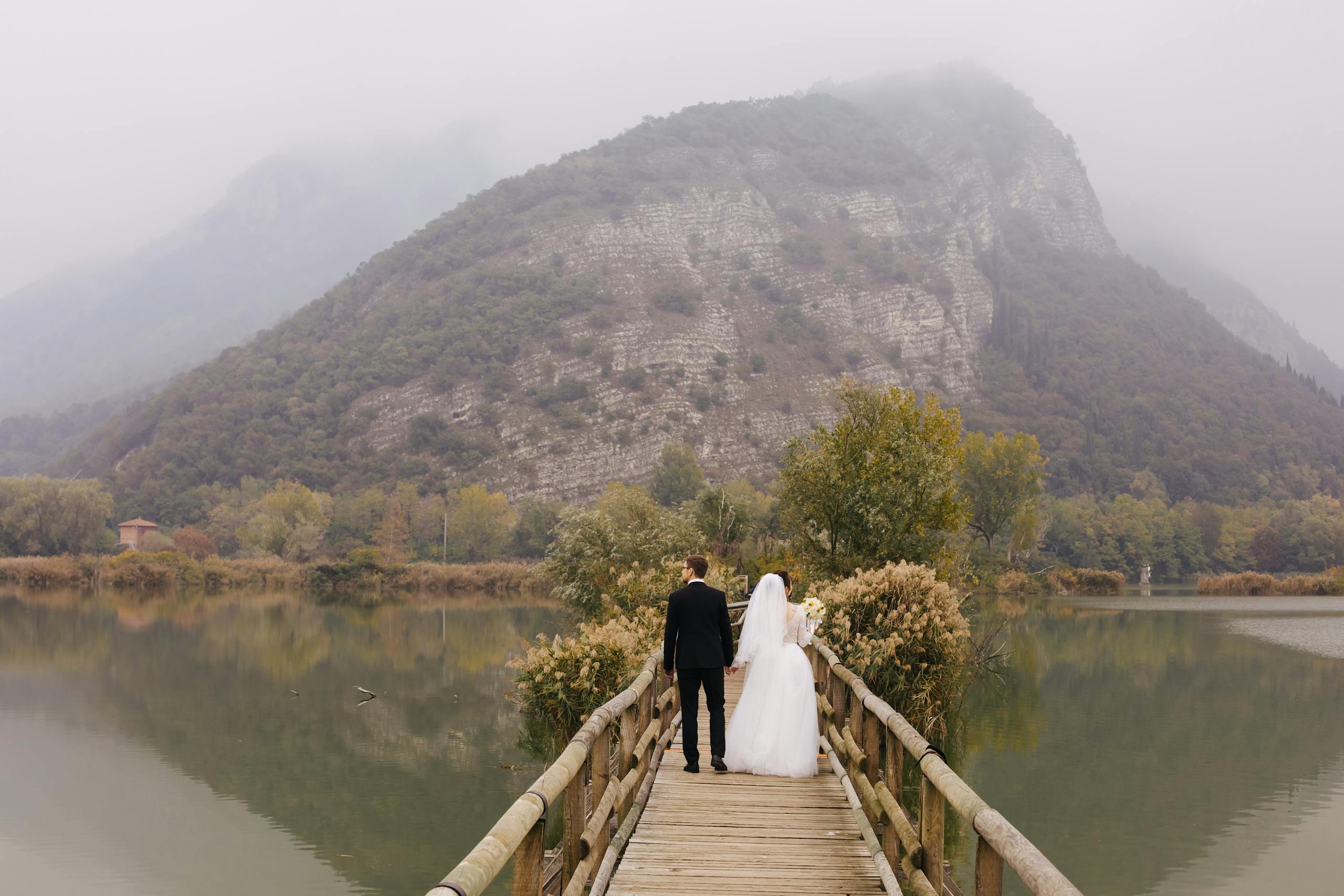 A newlywed couple walks hand in hand on a scenic bridge in Lombardy, Italy, surrounded by serene mountain views.