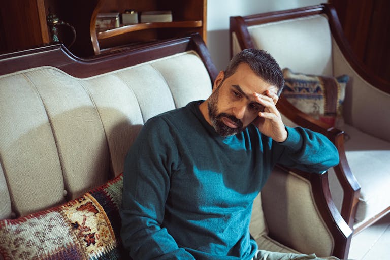 A pensive adult man in a teal sweater sits on a vintage sofa indoors, lost in thought.