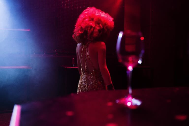 A performer in glamorous attire on a dimly lit club stage, with a wine glass in the foreground.