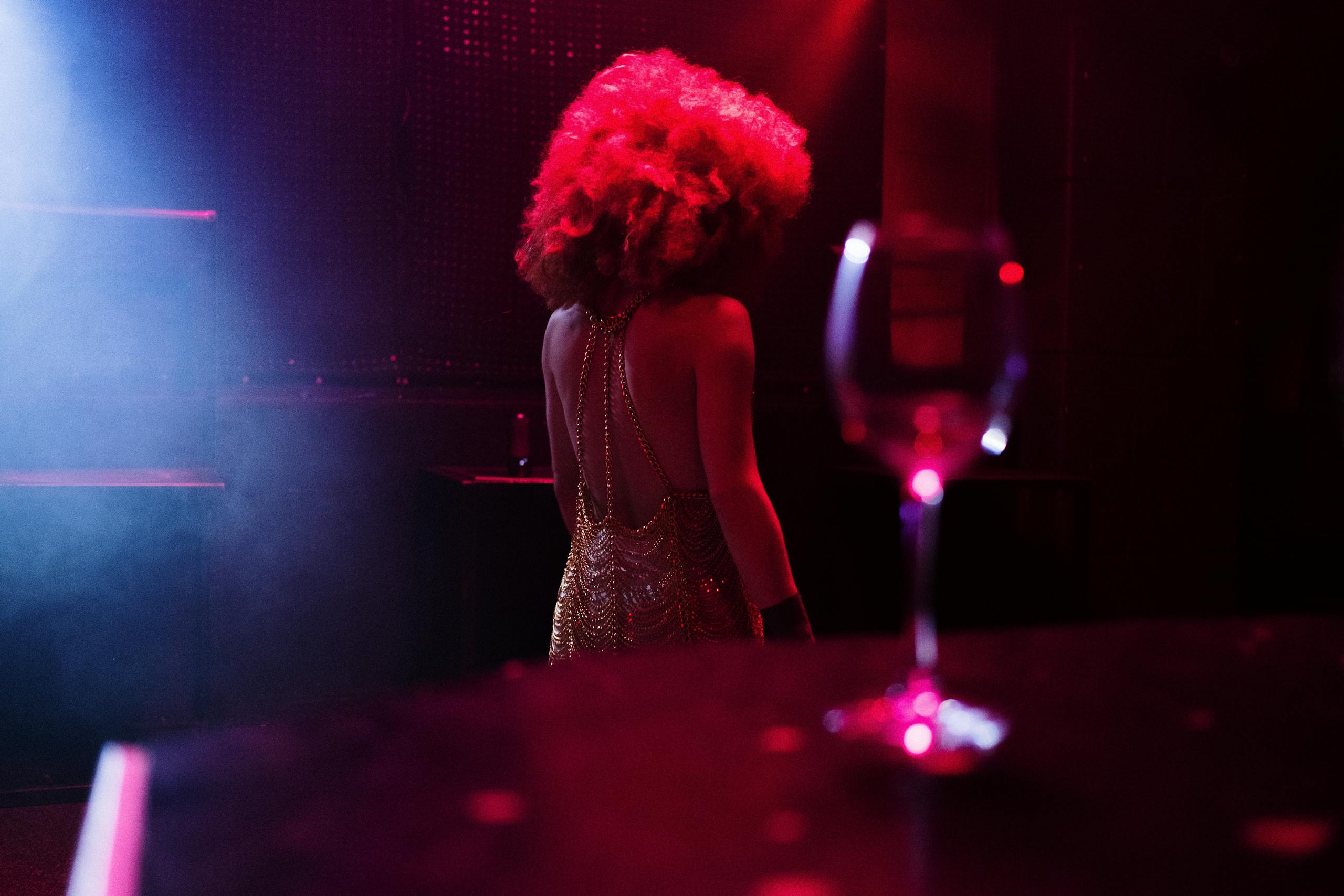 A performer in glamorous attire on a dimly lit club stage, with a wine glass in the foreground.