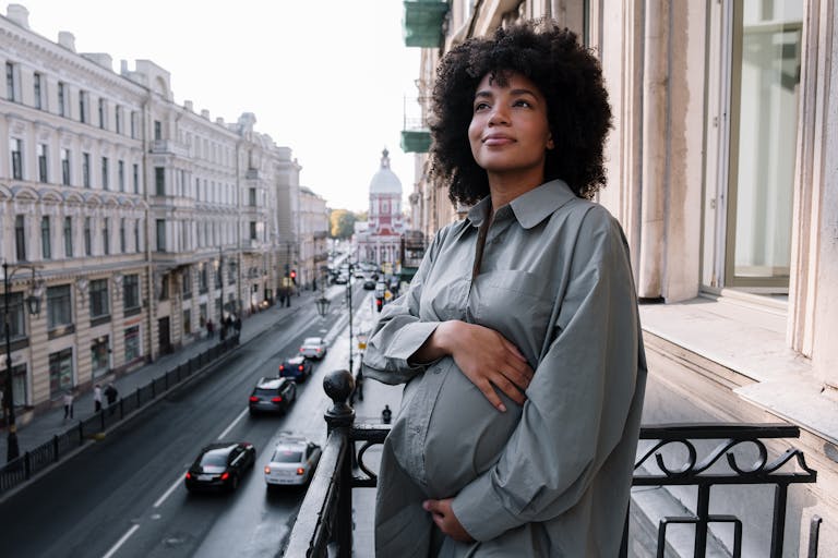 A pregnant woman stands on a balcony overlooking a bustling city street, radiating tranquility.