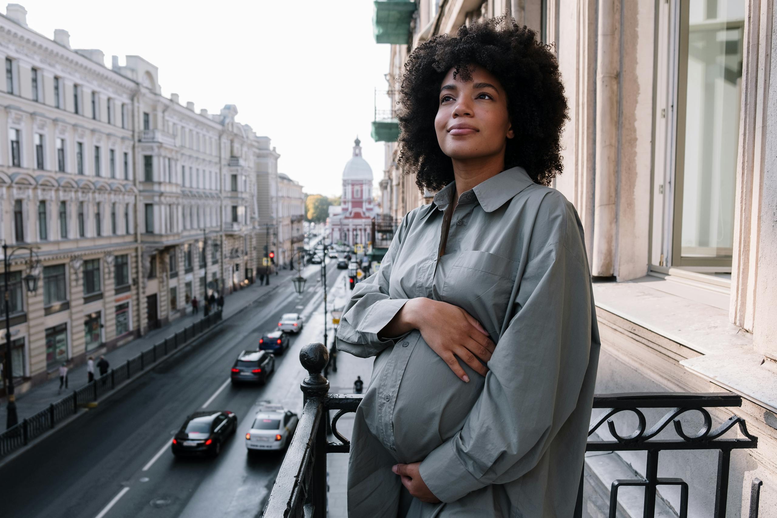 A pregnant woman stands on a balcony overlooking a bustling city street, radiating tranquility.