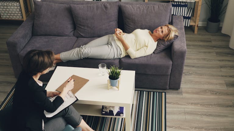 A therapist consulting with a patient reclining on a sofa in an office setting.