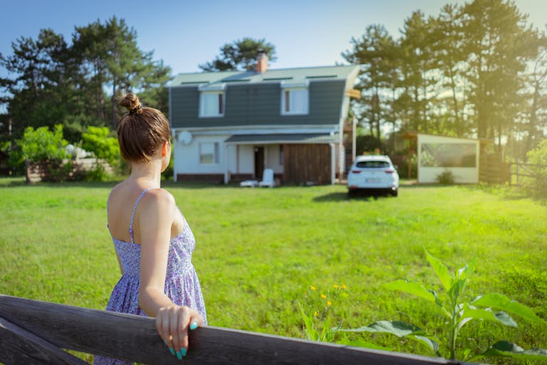 A woman in a sundress stands by a fence admiring a countryside house on a sunny day.