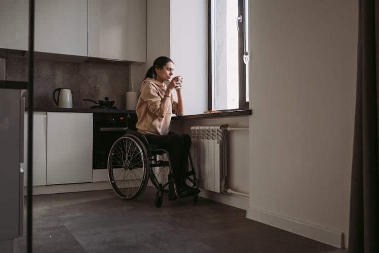 A woman in a wheelchair holding a coffee cup, sitting indoors by a window.