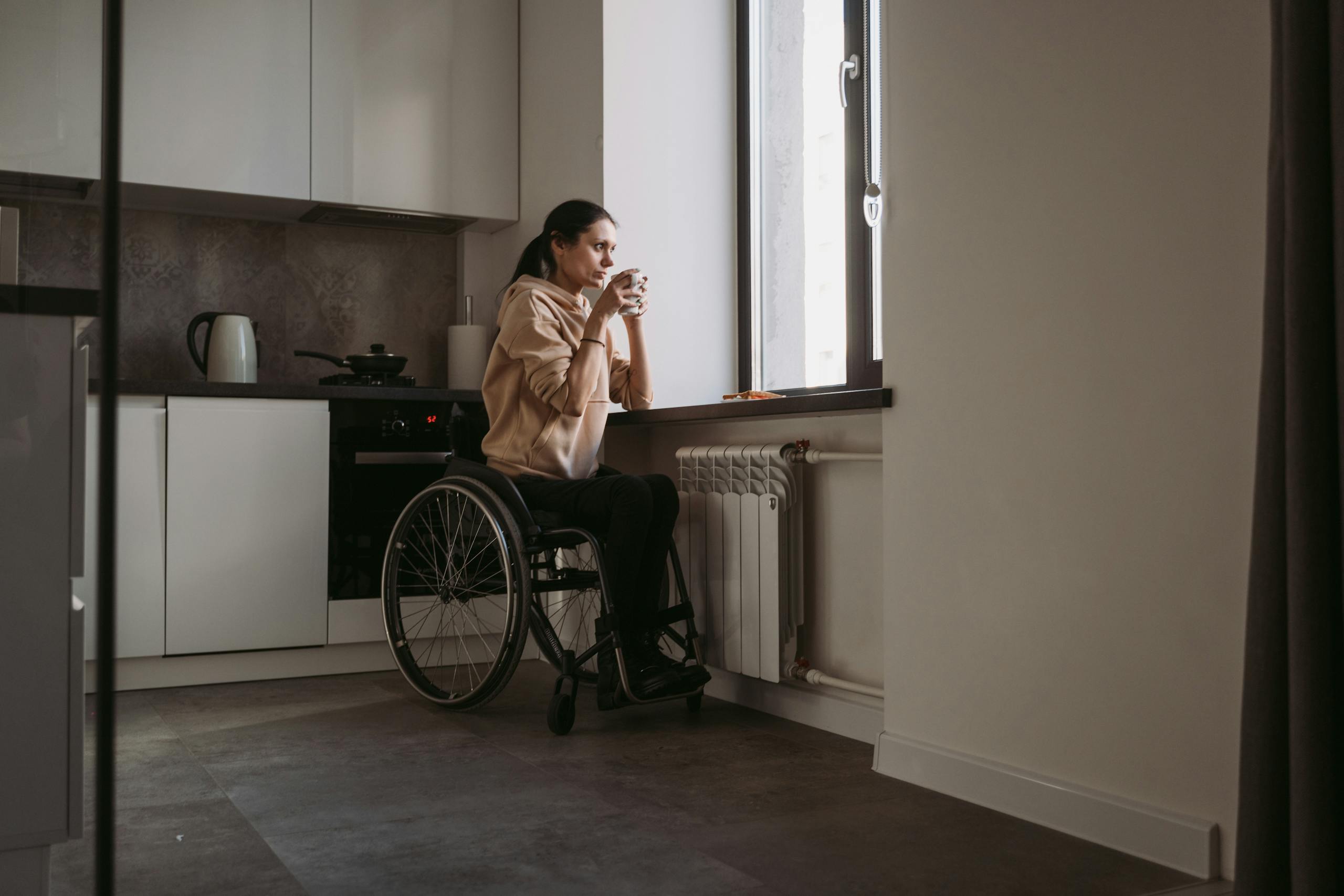 A woman in a wheelchair holding a coffee cup, sitting indoors by a window.