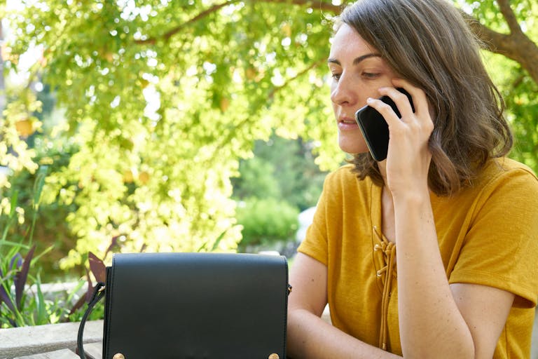 A woman in a yellow shirt makes a phone call outdoors amid lush greenery.