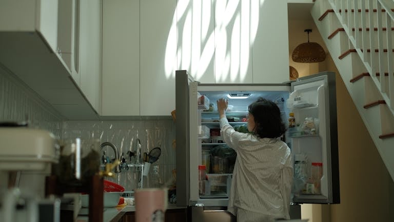 A woman in pajamas checks the refrigerator in a warmly lit cozy kitchen.