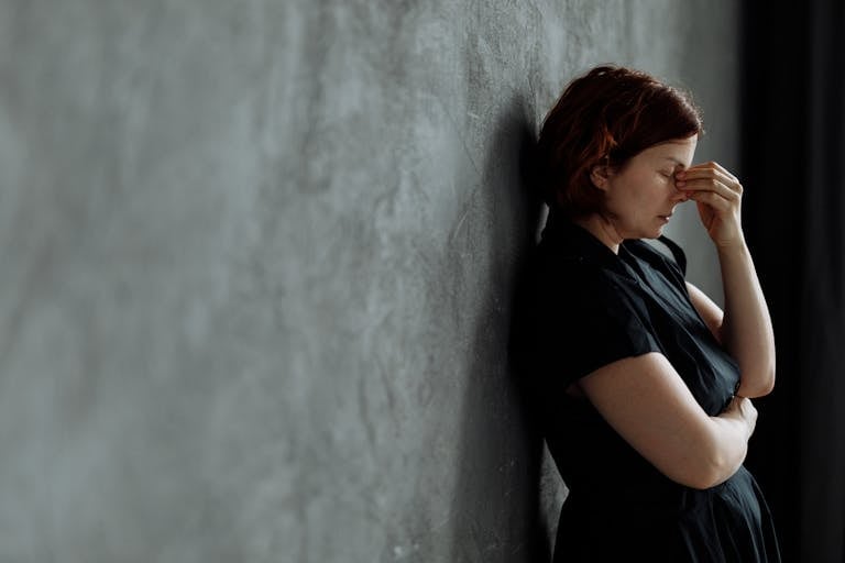 A woman in stress with eyes closed, leaning against a concrete wall indoors.