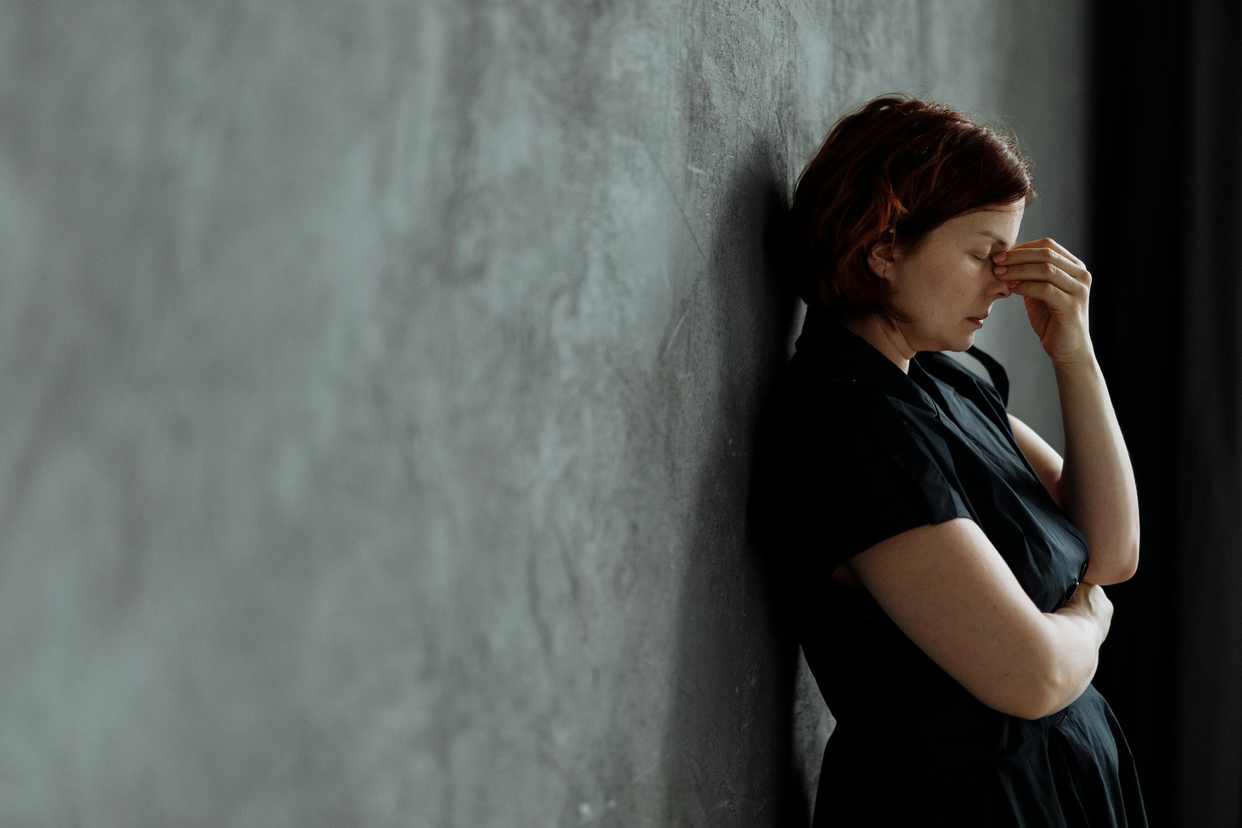 A woman in stress with eyes closed, leaning against a concrete wall indoors.