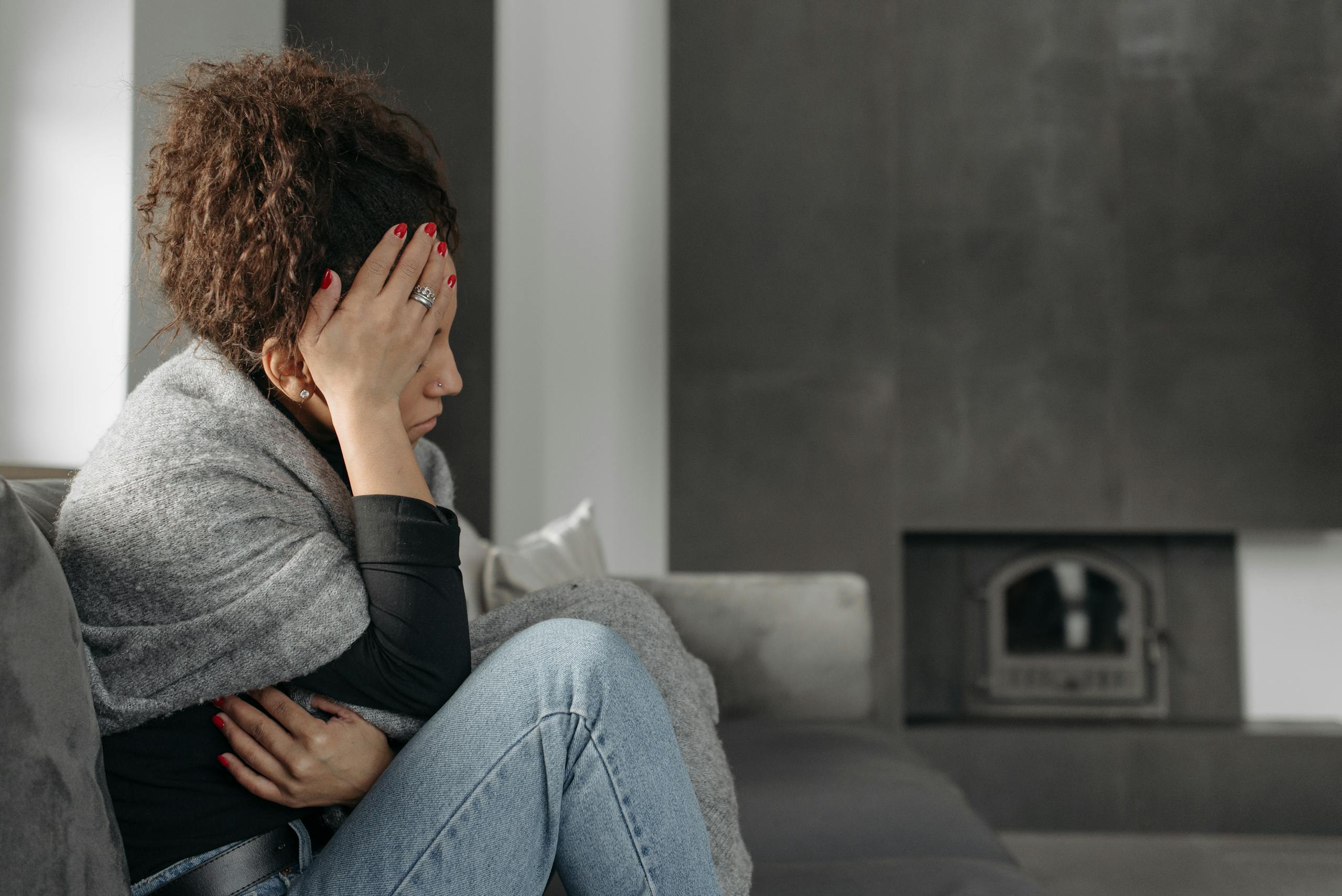 A woman sits on a couch indoors, wrapped in a blanket, holding her head in discomfort.