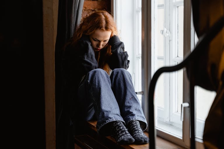 A woman sits pensively by a window, expressing feelings of solitude and contemplation.