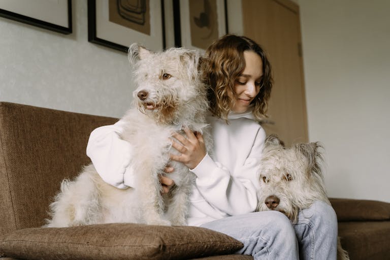 A woman sitting indoors, lovingly petting two fluffy dogs on a sofa.
