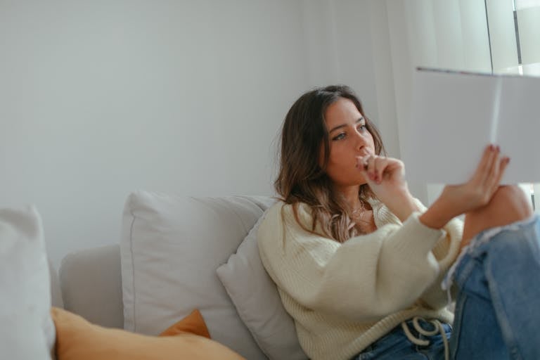 A woman sitting on a couch indoors, thoughtfully reading a book in a cozy setting.