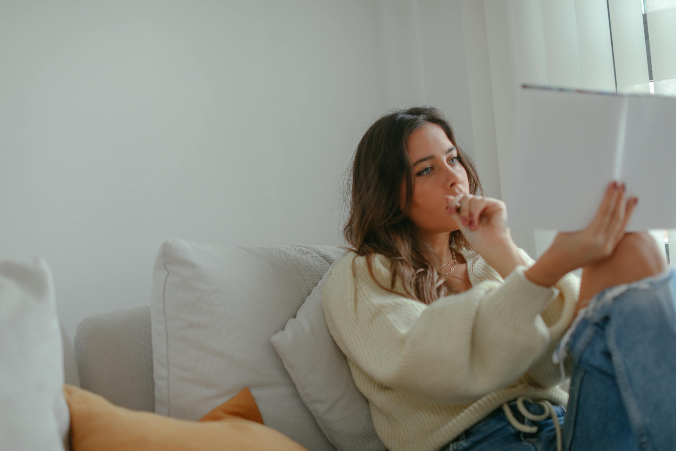 A woman sitting on a couch indoors, thoughtfully reading a book in a cozy setting.
