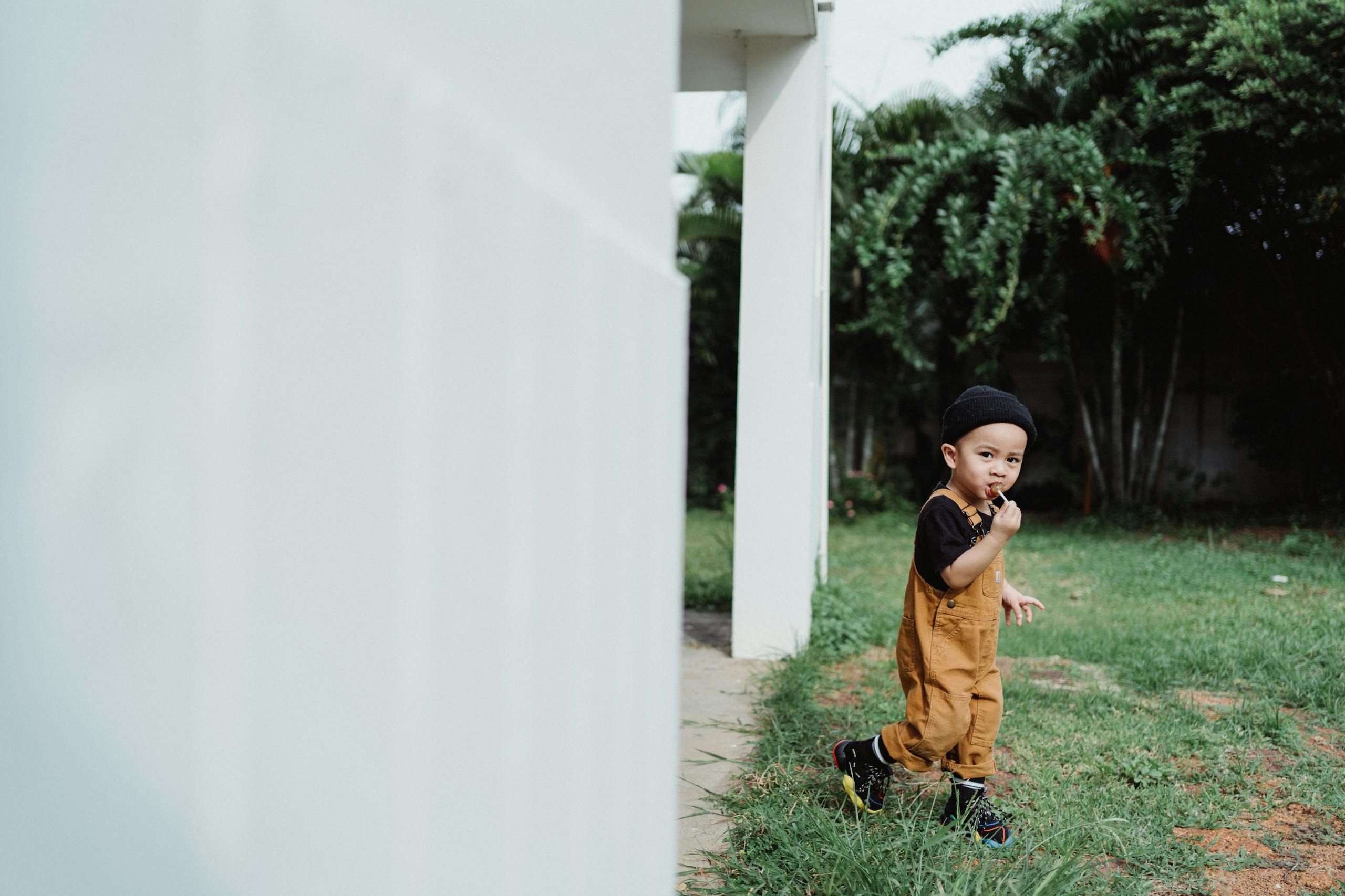 A young child in overalls enjoying a lollipop outdoors near a house.