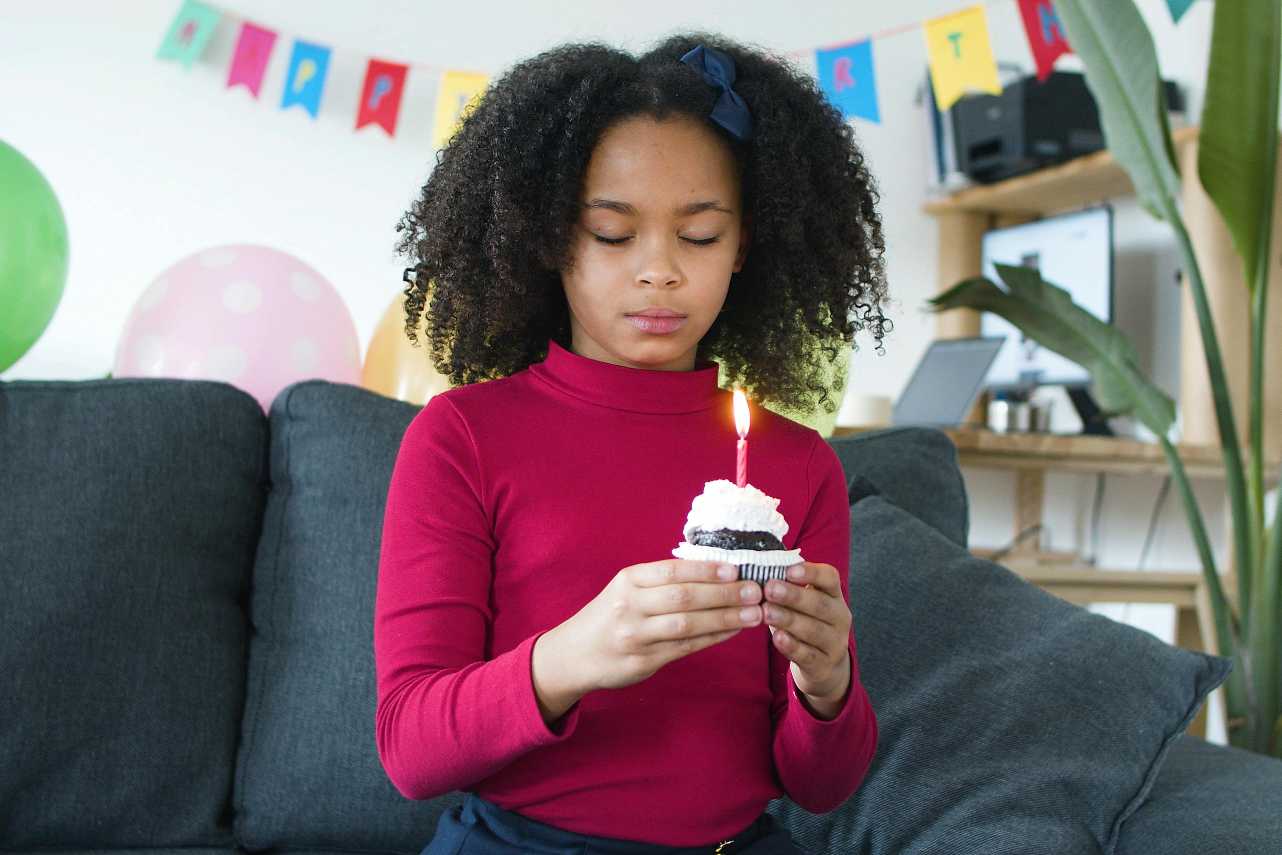 A young girl with curly hair holding a cupcake with a candle, eyes closed in a birthday setting.