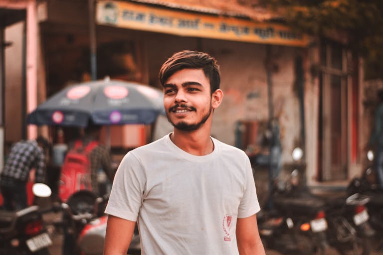 A young Indian man smiling outdoors in casual white attire on a bustling street.