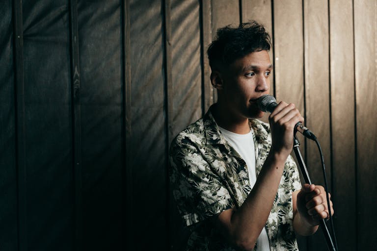 A young male singer in a floral shirt performing indoors with a microphone.
