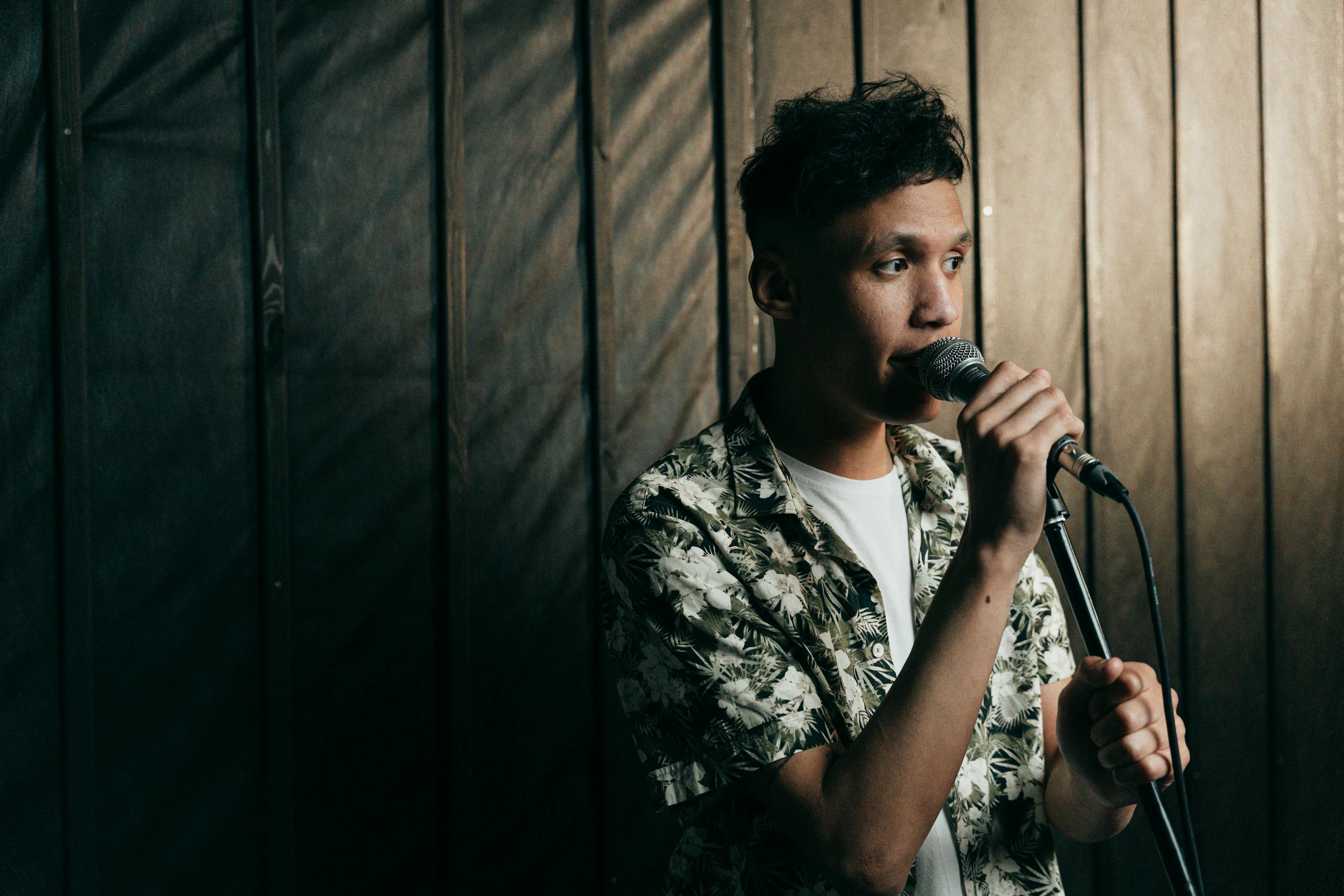 A young male singer in a floral shirt performing indoors with a microphone.