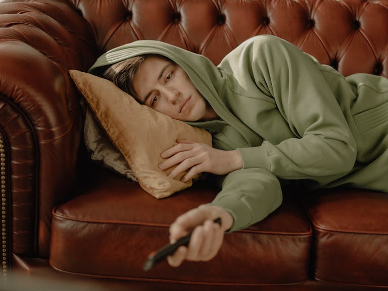 A young man in a green hoodie relaxes on a leather sofa with a remote control, depicting a cozy home scene.