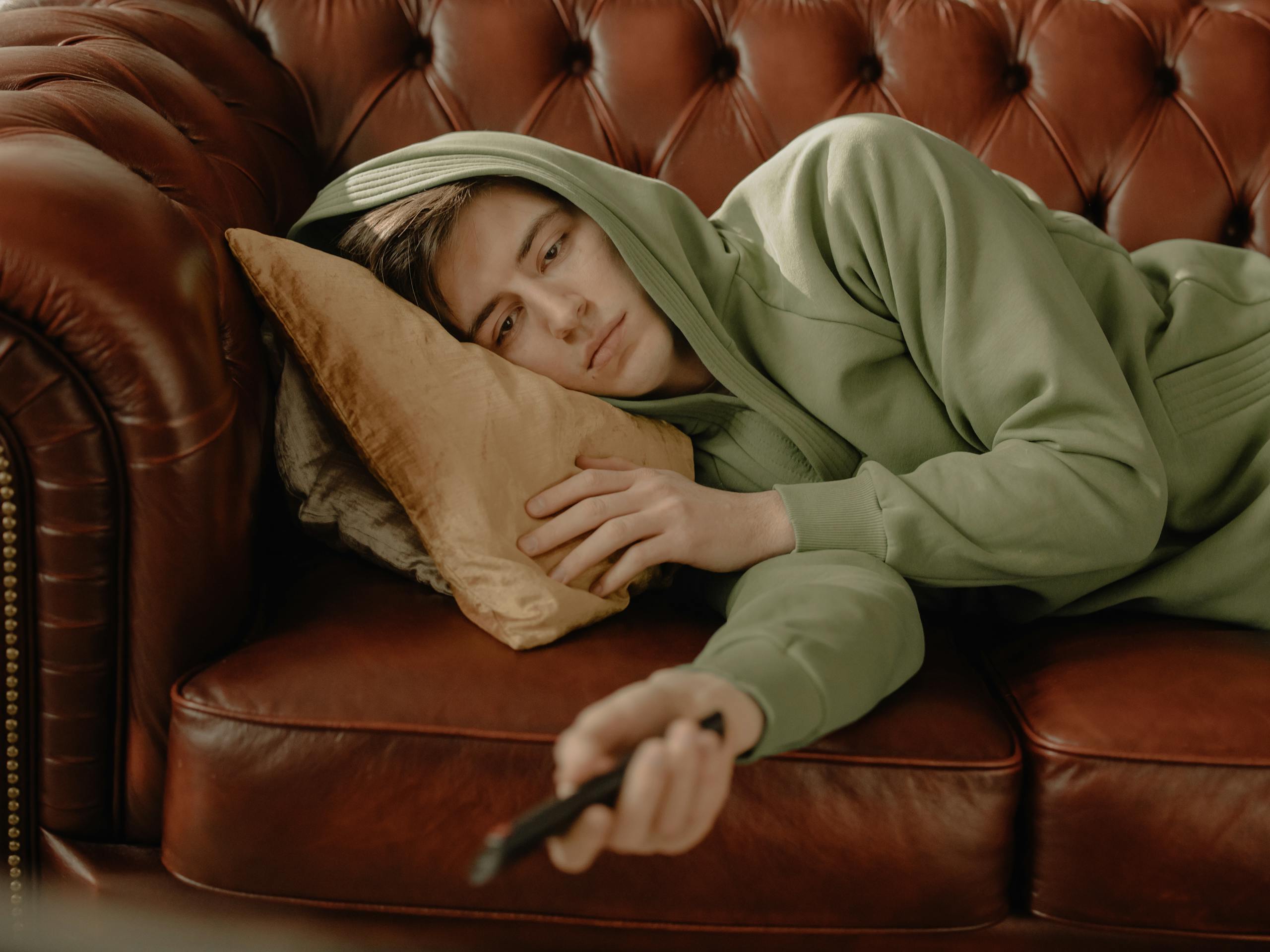 A young man in a green hoodie relaxes on a leather sofa with a remote control, depicting a cozy home scene.