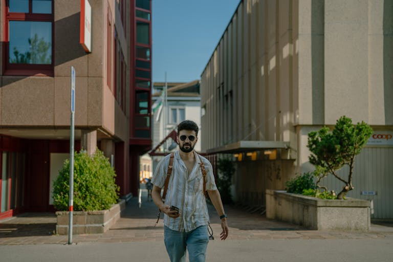 A young man with sunglasses and a backpack walks down an urban street, embracing a modern lifestyle.