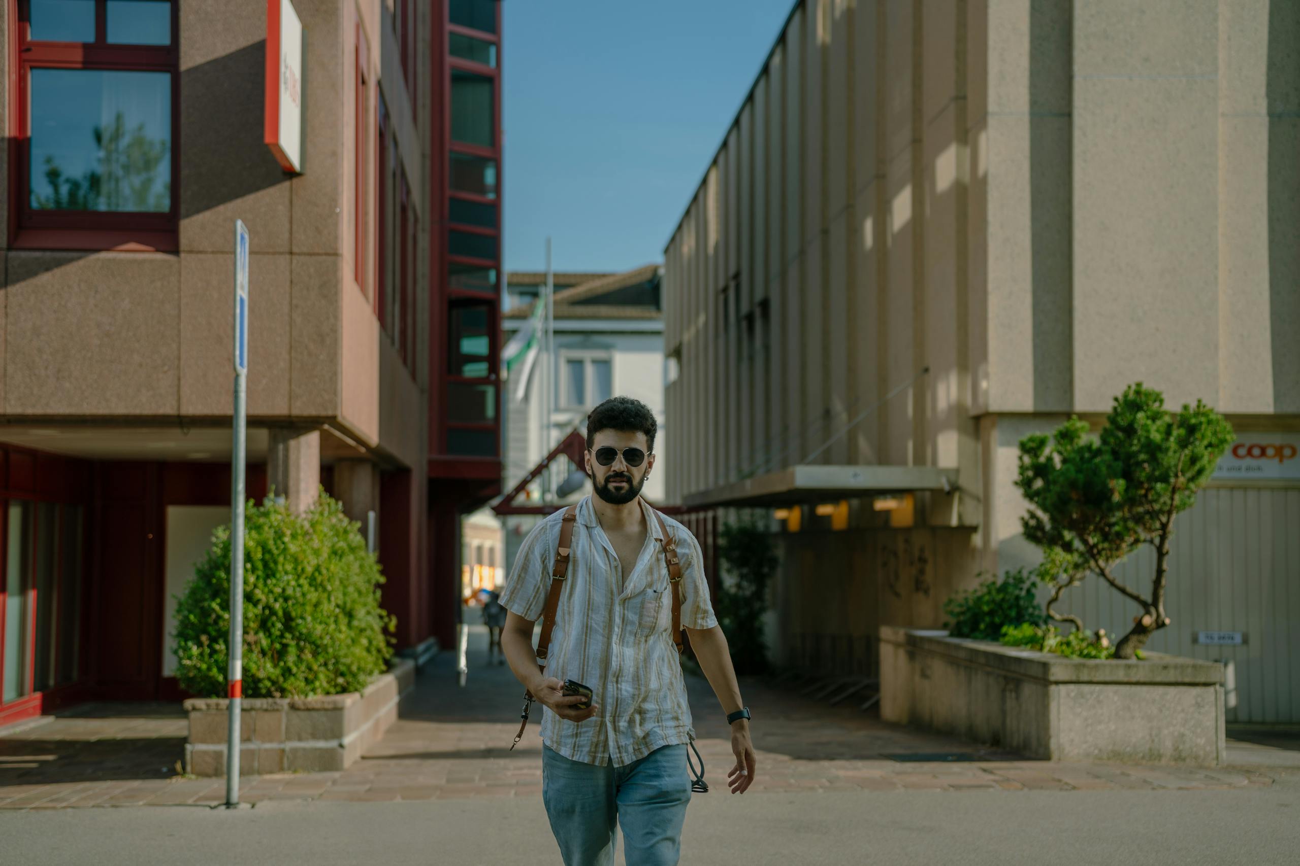 A young man with sunglasses and a backpack walks down an urban street, embracing a modern lifestyle.