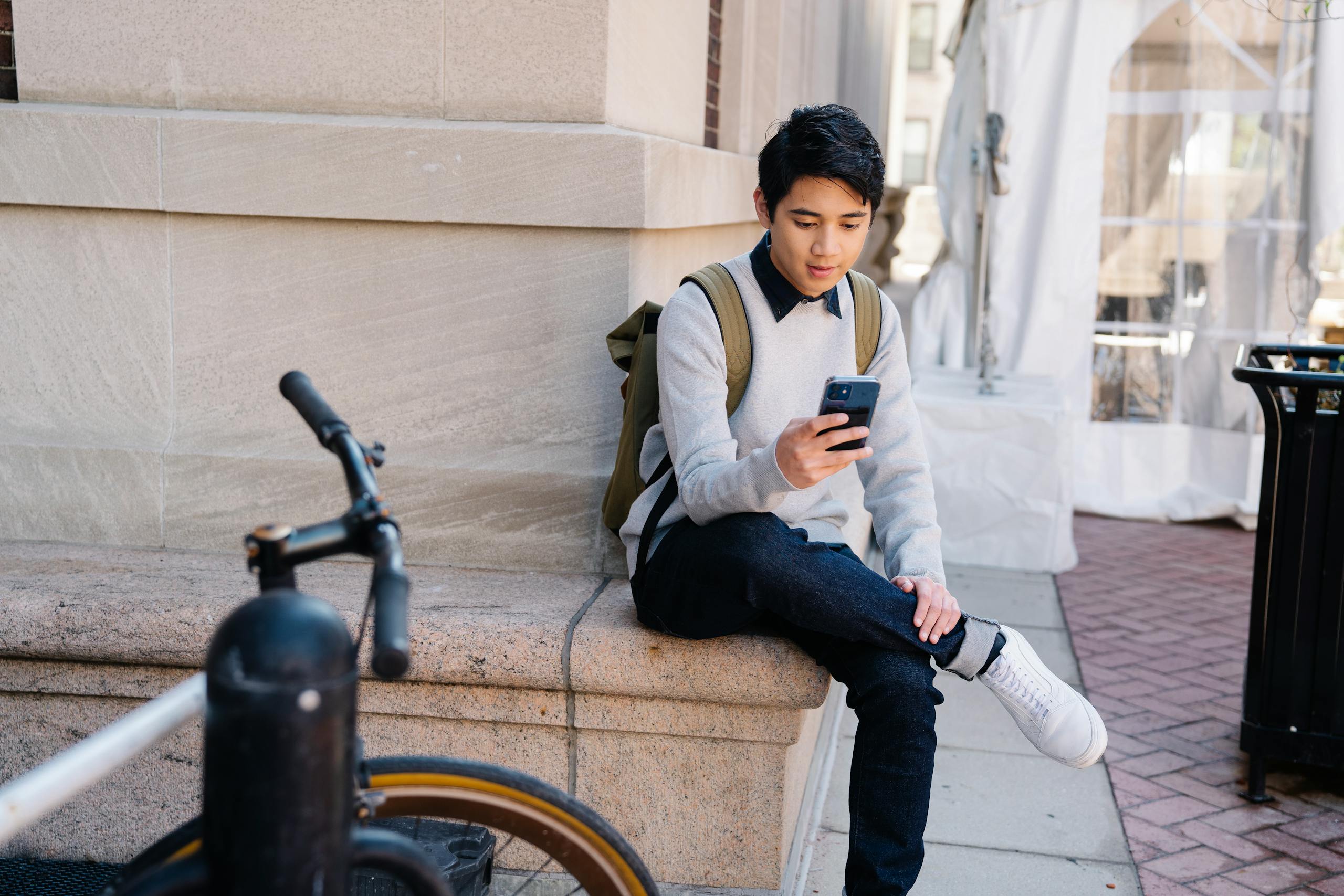 A young student in casual attire uses a smartphone while sitting outdoors on a university campus.