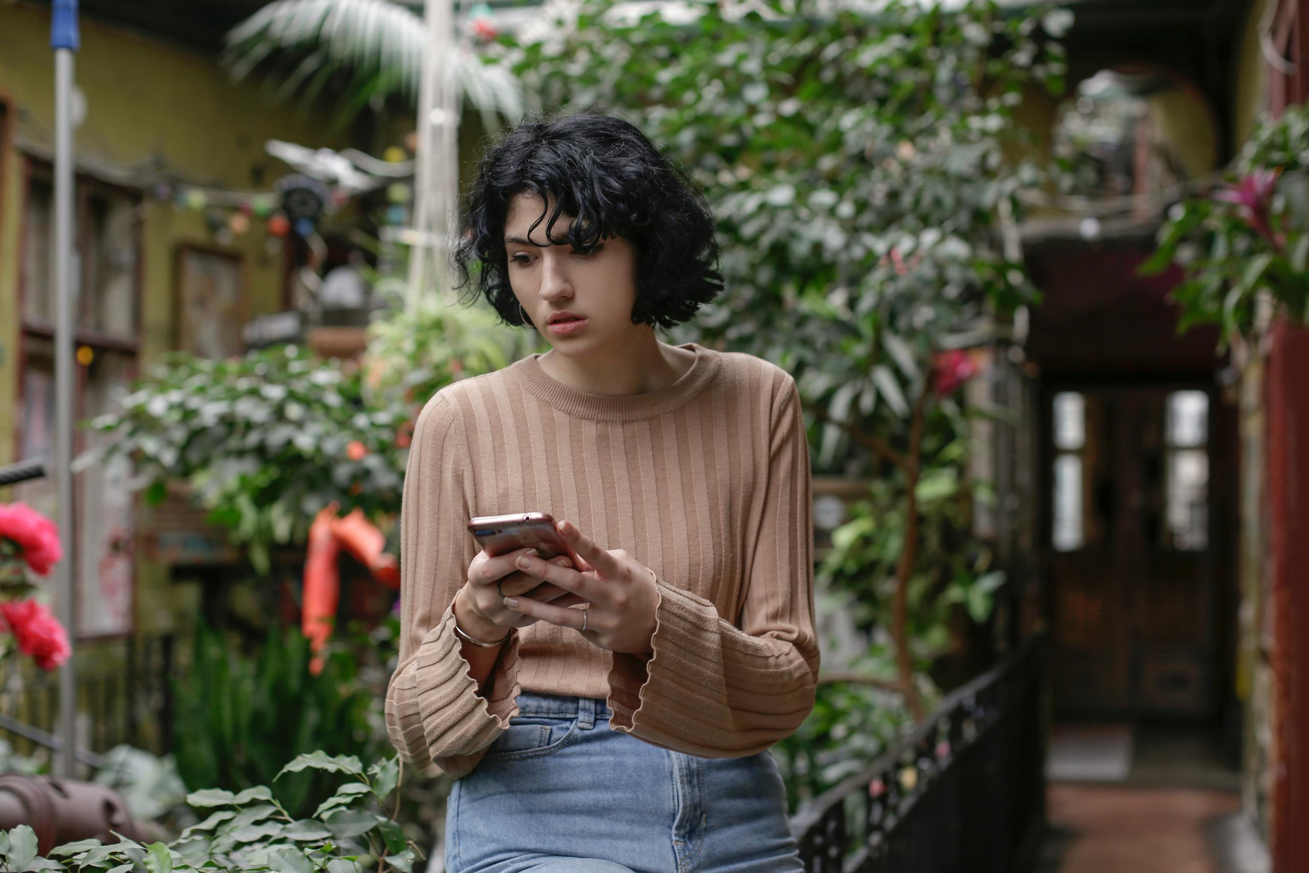 A young woman browsing her smartphone in a lush indoor garden with vibrant greenery.