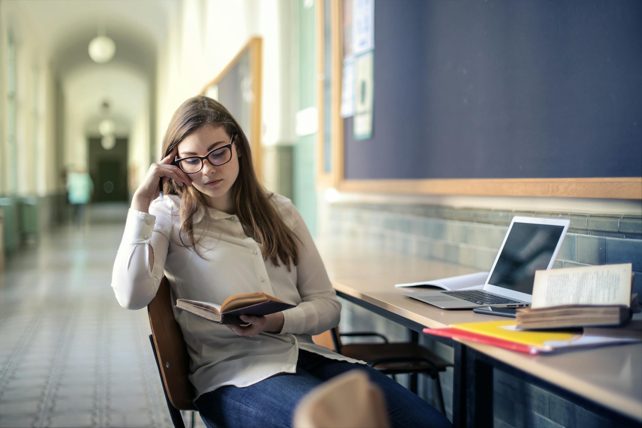 A young woman studies diligently in a university hallway, reading a book.