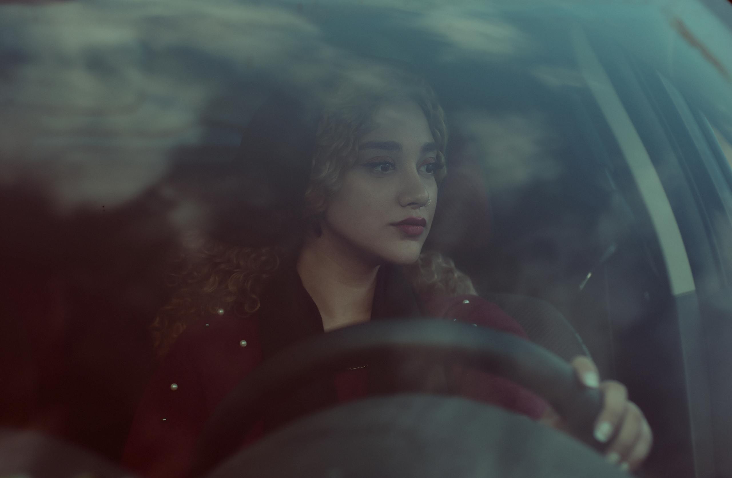 A young woman with curly hair focuses on driving through the city, reflecting her thoughtful expression.