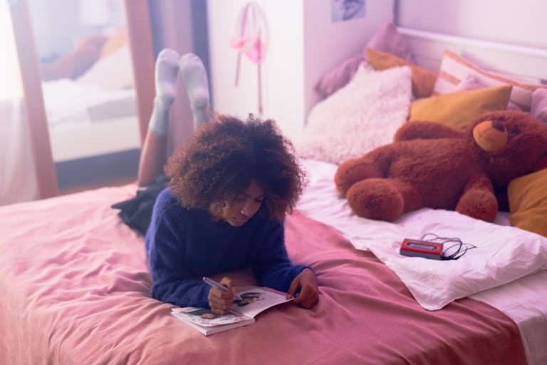 A young woman writing in her journal, lying on a bed with a teddy bear nearby, creating a cozy indoor scene.
