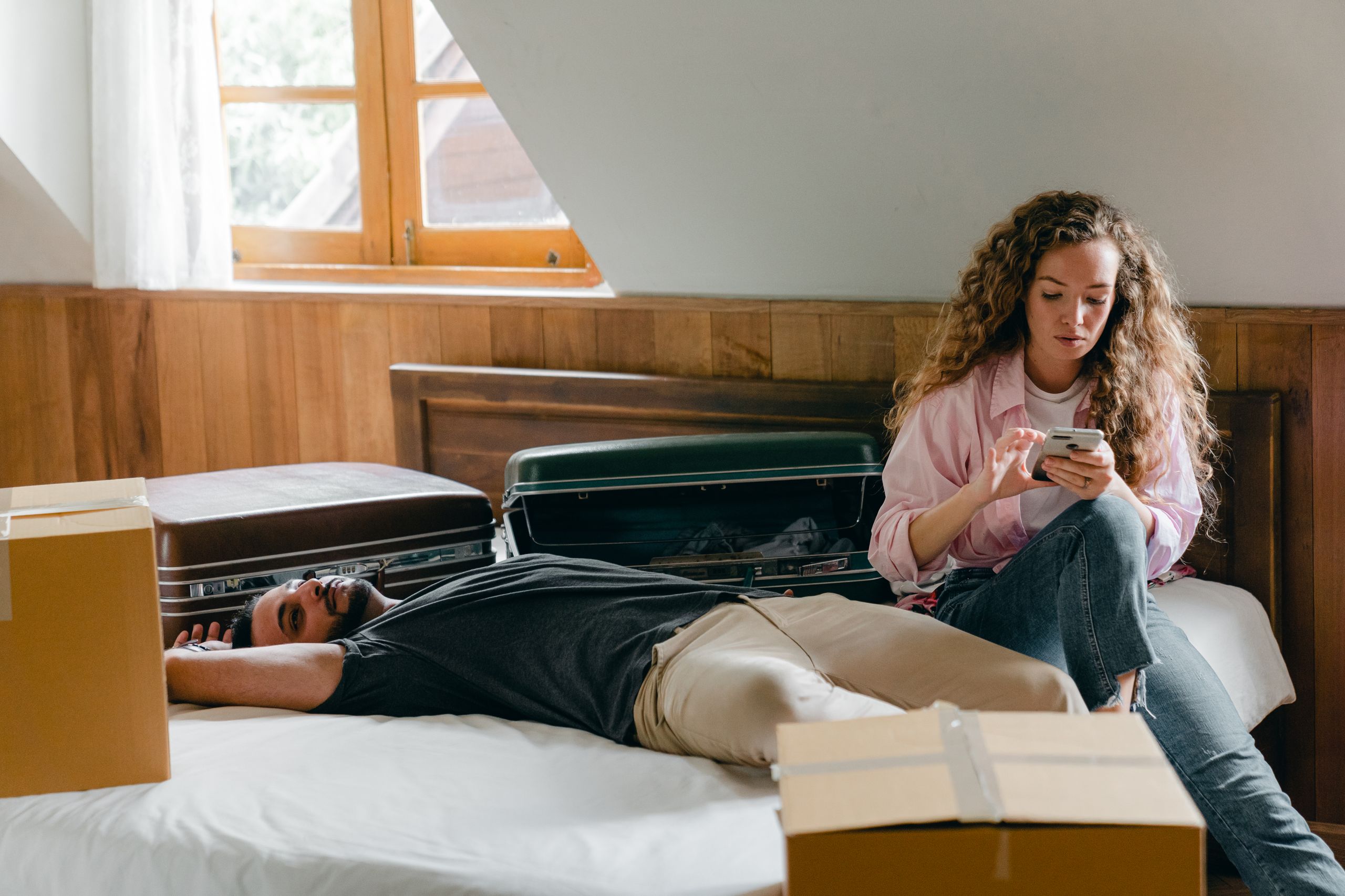 Adult ethnic man lying on bed while woman with curly hair using smartphone sitting near and relaxing among suitcases and cardboard boxes for moving