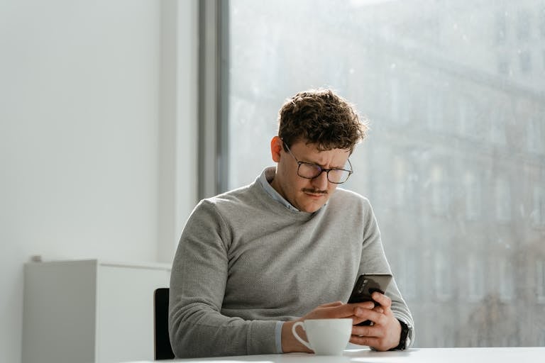 Adult man wearing glasses using smartphone in sunny office space, holding a mug.