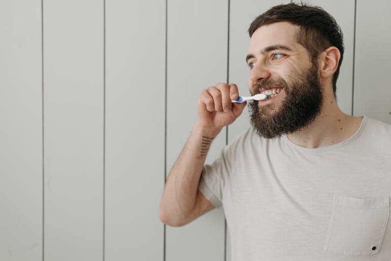Adult man with a beard brushing his teeth indoors with a manual toothbrush.