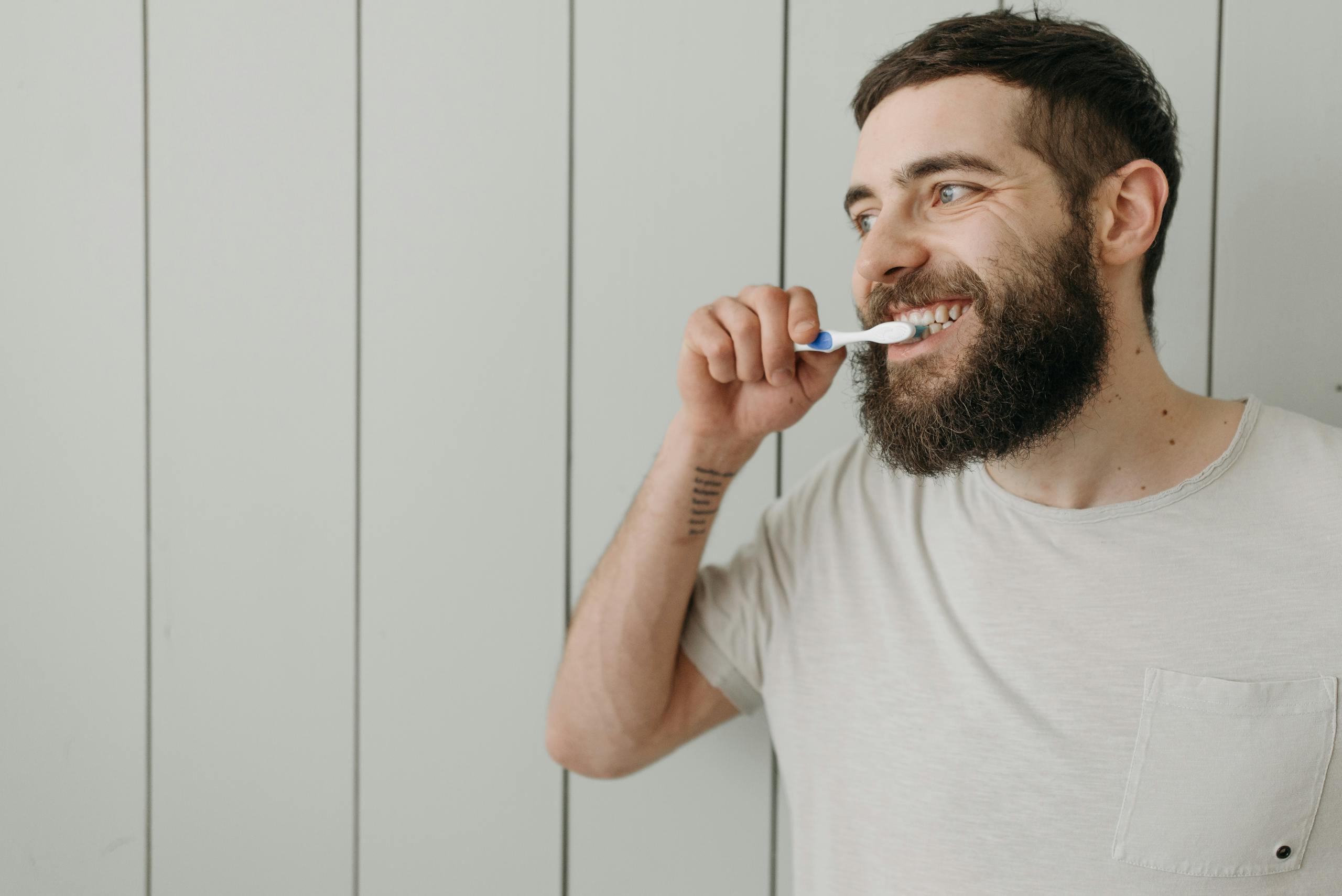 Adult man with a beard brushing his teeth indoors with a manual toothbrush.