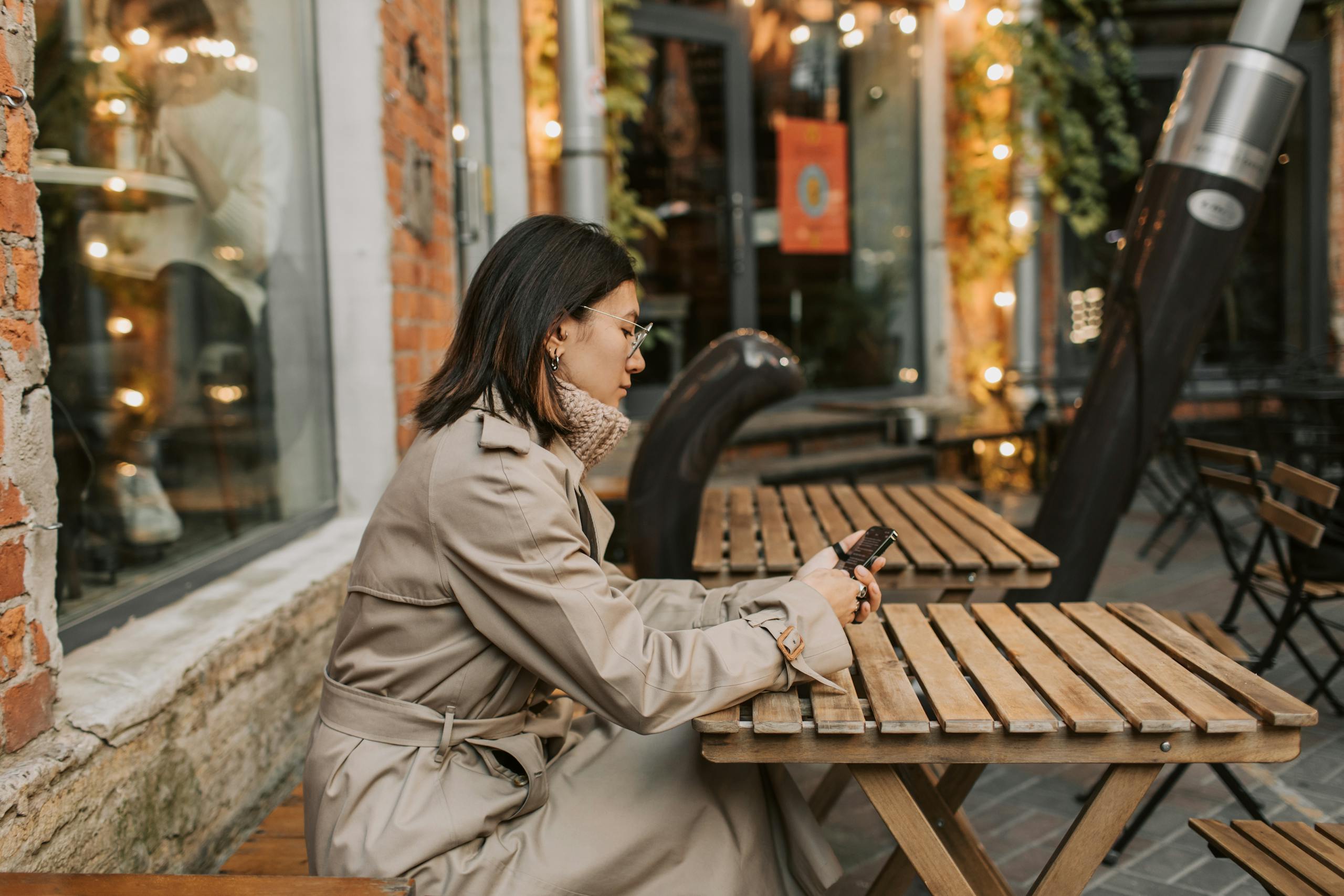 Asian woman in trench coat browsing smartphone at an outdoor café wooden table.