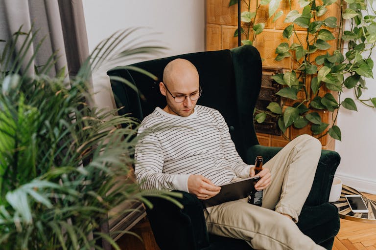 Bald man in glasses relaxing in an armchair while reading on a tablet in a plant-filled living room.