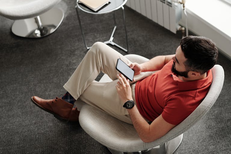 Bearded man in a red polo shirt sitting comfortably, using a smartphone indoors.