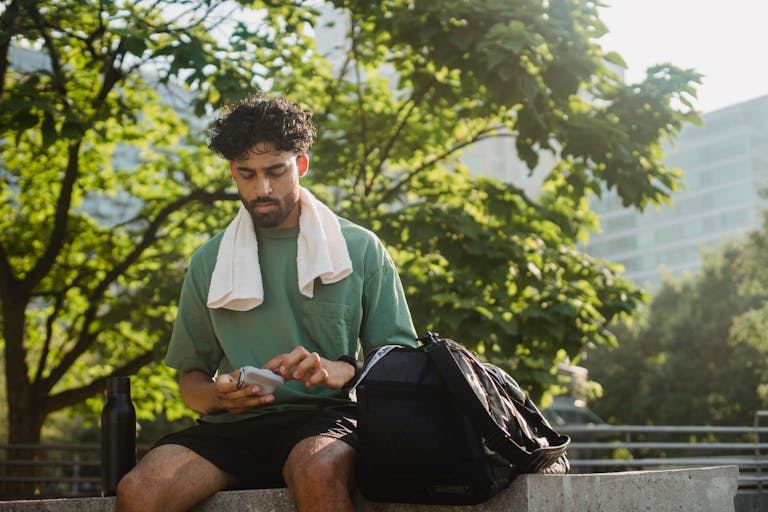Bearded man sitting outdoors, checking phone, relaxing on a sunny day.