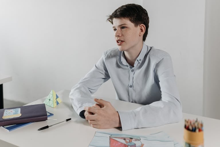 Boy sitting at a desk in a classroom, surrounded by books and drawings, looking thoughtful and engaged.