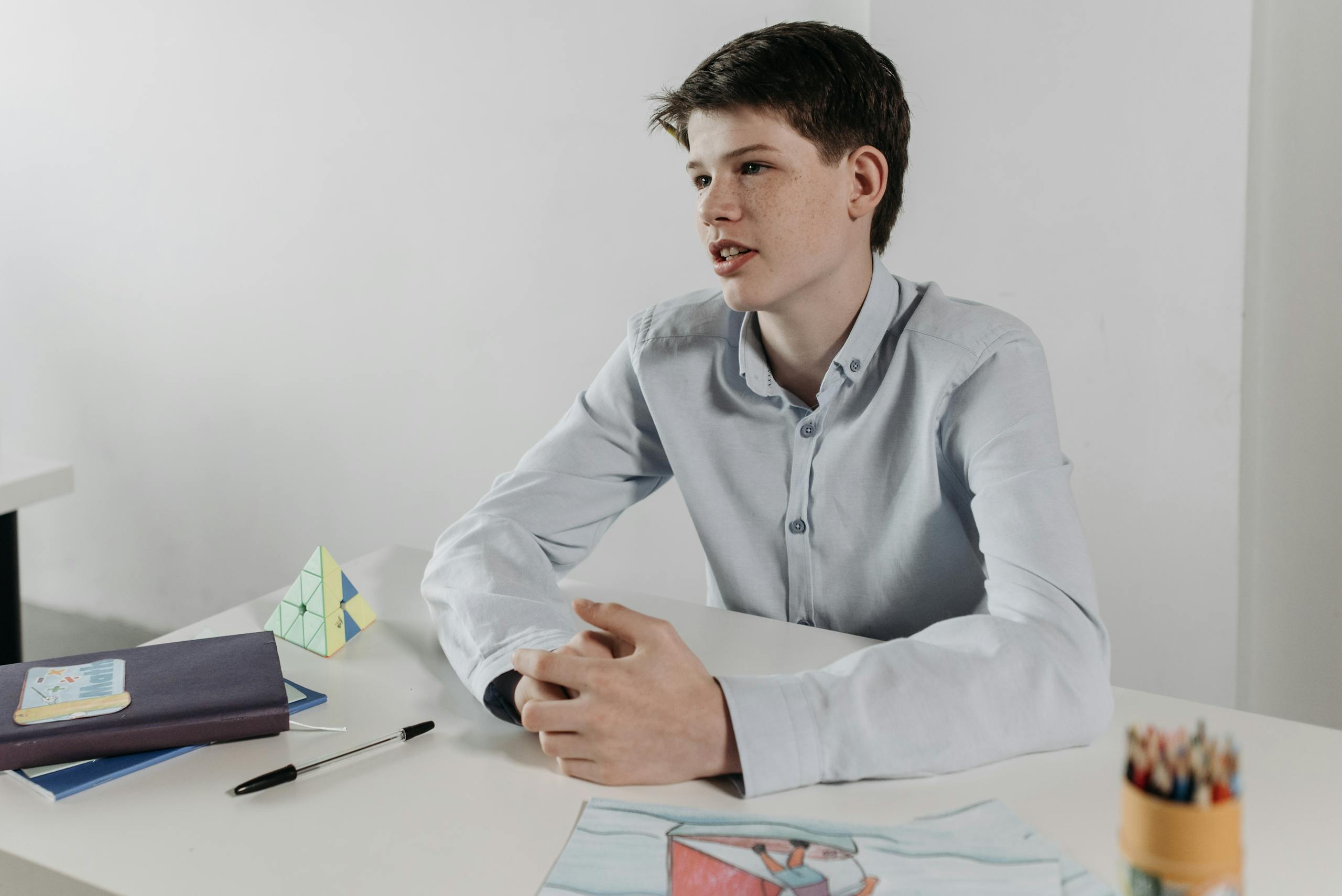 Boy sitting at a desk in a classroom, surrounded by books and drawings, looking thoughtful and engaged.