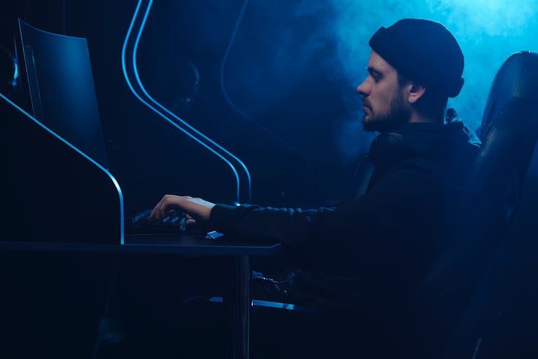Caucasian man engrossed in gaming on a computer in a dark, moody setting.