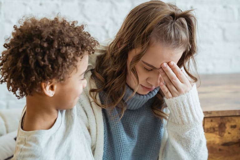 Cheerful African American son with curly hair consoling unhappy mother with closed eyes sitting in light room at home near white wall